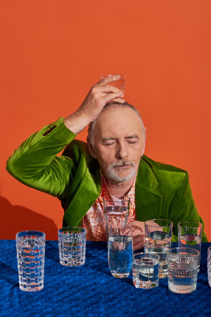 bearded senior man with closed eyes, in stylish and green velvet blazer, holding glass of water above head on orange background, aging population, symbolism, life fullness conceptの写真素材