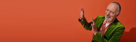 happy and trendy aging concept, excited grey haired and bearded senior man in green velour blazer showing stop gesture and laughing with closed eyes on red orange background, banner with copy spaceの写真素材