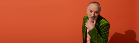 curious and joyful senior man with grey hair and beard holding hand near chin and looking at camera on red orange background, older model, green velour blazer, positive and stylish aging, bannerの写真素材