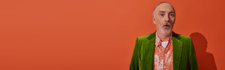 trendy senior man with shocked face expression looking at camera on red orange background, grey hair and beard, green velour blazer, fashion and age concept, banner with copy spaceの写真素材