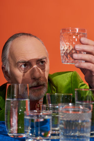 senior and thoughtful grey haired man in green velvet blazer looking at glass full of pure water...