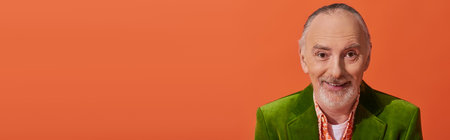 portrait of trendy and cheerful senior model with grey hair and beard, wearing green velour blazer and smiling at camera on vibrant orange background, fashion and age concept, banner with copy spaceの写真素材