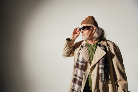 elderly man in beanie hat, beige trench coat and plaid scarf adjusting dark sunglasses and looking away on grey background, hipster style, individuality, fashion and age conceptの写真素材