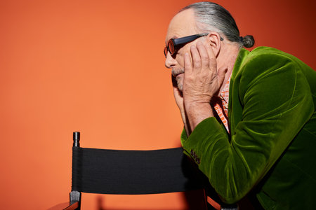side view of senior grey haired model touching face and looking away near chair on red and orange background, fashion look, dark sunglasses, green velour blazer, fashionable aging conceptの写真素材
