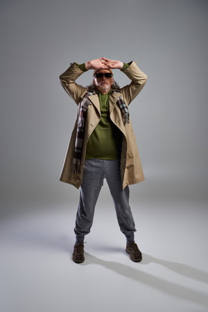 full length of cool and senior hipster man in dark sunglasses, beanie hat, beige trench coat and sneakers posing with hands above head on grey background, fashionable aging conceptの写真素材