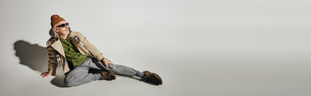 full length of elderly man in dark sunglasses, beanie hat, beige trench coat and sneakers sitting and looking away on grey background with shadow, hipster fashion, banner with copy spaceの写真素材