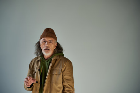 frustrated and worried elderly man with grey hair and beard, in beanie hat and brown jacket looking...