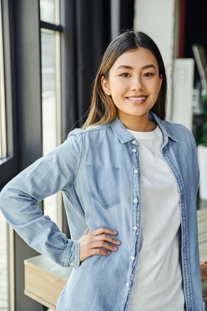 young and attractive asian businesswoman in blue denim shirt, with brunette hair and radiant smile standing with hand on hip and looking at camera in modern office, positive emotionの写真素材