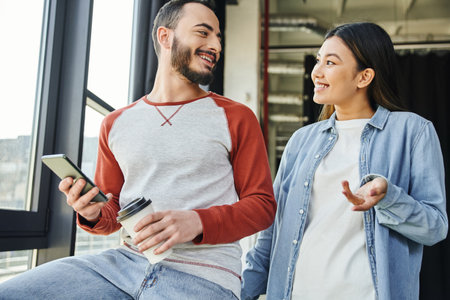 happy asian woman in stylish casual clothes talking to bearded colleague holding smartphone and paper cup with takeaway drink in modern office, young interracial entrepreneurs planning startupの写真素材