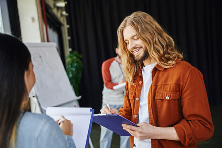 stylish and long haired man smiling while writing on clipboard near woman and medical instructor with flip chart on blurred background, first aid seminar, health care and emergency response conceptの写真素材