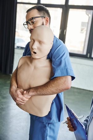 first aid seminar, young professional paramedic showing rescue techniques in case of chocking on CPR manikin near woman with clipboard and pen in training room, emergency situations preparednessの写真素材