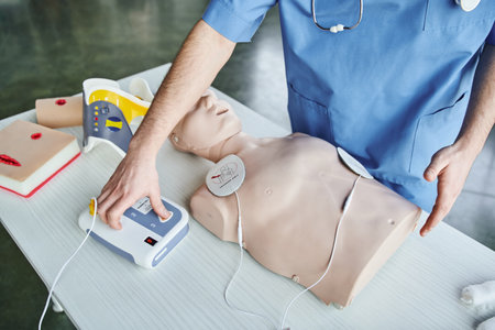 partial view of medical instructor operating automated defibrillator near CPR manikin, wound care simulators and neck brace, first aid hands-on learning and critical skills development conceptの写真素材