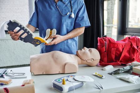 partial view of healthcare worker in blue uniform holding neck brace near CPR manikin,...