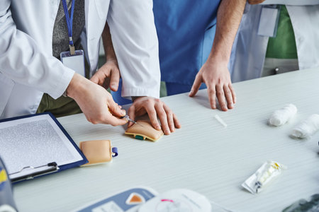 partial view of student in white coat doing injection in training pad near clipboard, bandages and...
