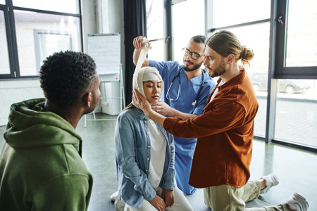 young man and medical instructor bandaging head of asian woman near african american participant of first aid seminar in training room, importance of emergency preparedness conceptの写真素材
