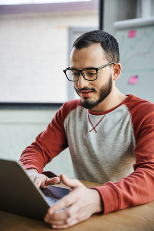 positive and concentrated bearded entrepreneur in eyeglasses and casual clothes sitting at workplace...