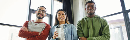 joyful asian woman with coffee to go and multiethnic businessmen with folded arms looking at camera in modern office, creative team, successful collaboration, bannerの写真素材