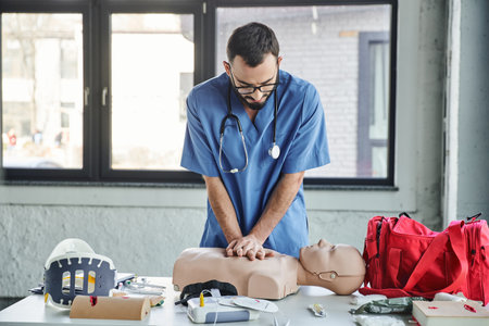 young paramedic in blue uniform and eyeglasses practicing chest compressions on CPR manikin near defibrillator and first aid kit during medical seminar, life-saving skills development conceptの写真素材