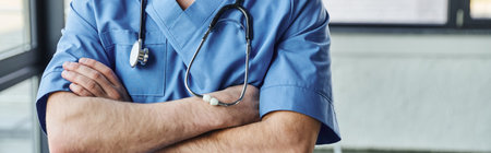 partial view of young doctor in blue uniform standing with crossed arms and stethoscope on neck in modern hospital, first aid training seminar and emergency preparedness concept, bannerの写真素材