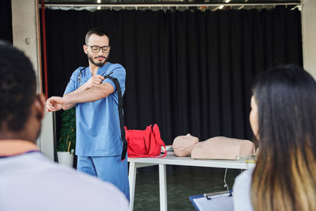 first aid seminar, medical instructor in blue uniform and eyeglasses applying compression tourniquet near interracial participants on blurred foreground, acquiring and practicing life-saving skills conceptの写真素材