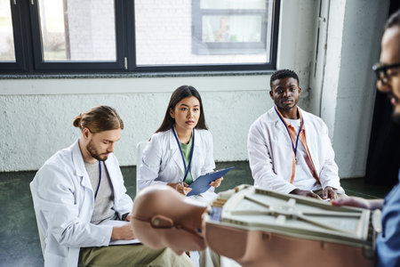 medical training, diverse group of multiethnic students in white coats looking at paramedic standing with CPR manikin on blurred foreground, acquiring and practicing life-saving skills conceptの写真素材
