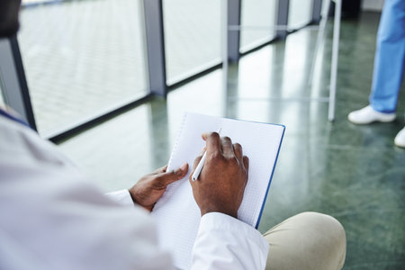 cropped view of african american student in white coat sitting with notebook and pen near medical instructor on blurred background, emergency preparedness and life-saving skills development conceptの写真素材