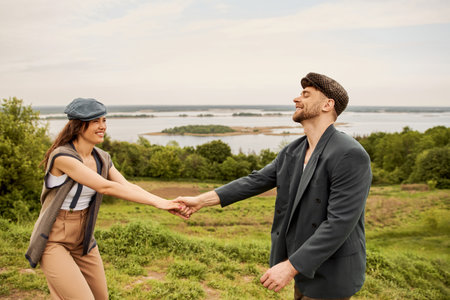 Positive and fashionable brunette woman in newsboy cap and vest holding hand of bearded boyfriend in jacket while standing with scenic landscape at background, fashionable couple in countrysideの写真素材