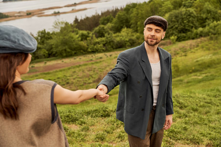 Smiling bearded man in newsboy cap and jacket holding hand of brunette girlfriend in vest while standing with nature at background, fashionable couple in countrysideの写真素材