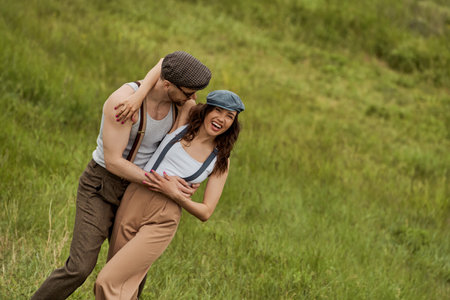 Fashionable man in vintage outfit and sunglasses hugging cheerful girlfriend in suspenders and newsboy cap while spending time together and standing on grassy lawn, stylish pair amidst natureの写真素材