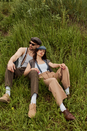 Trendy romantic couple in vintage outfits and newsboy caps relaxing next to each other while sitting...
