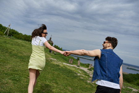 Side view of positive romantic couple in sunglasses and summer outfits holding hands while running...