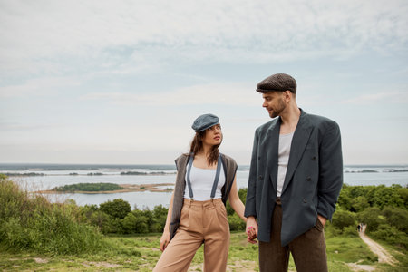 Fashionable and bearded man in newsboy cap and jacket holding hand of brunette girlfriend in suspenders and vest and standing together with nature at background, stylish pair amidst natureの写真素材