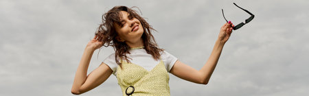 Low angle view of positive brunette woman in stylish sundress touching hair, looking at camera and...