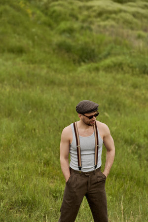 Trendy man in vintage-inspired outfit sunglasses and suspenders holding hands in pockets of pants while standing on blurred grassy meadow at background, man enjoying country lifeの写真素材
