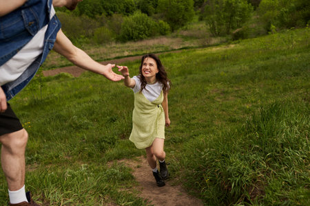 Cheerful brunette woman in stylish sundress and boots holding hand of boyfriend while walking on grassy hill at summer, rural tranquility and relaxing in nature conceptの写真素材