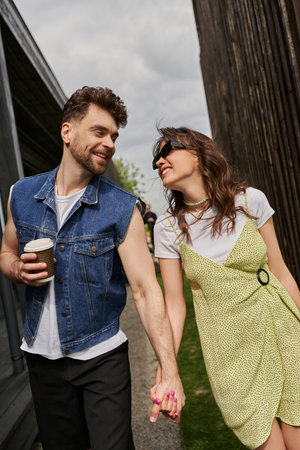 Cheerful romantic couple in stylish summer outfits holding hands and coffee to go while talking and...