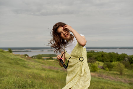 Positive brunette woman in trendy sundress holding sunglasses and touching hair while standing and...