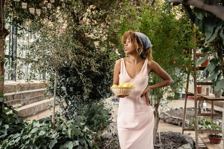 Trendy young african american woman in summer outfit holding hand on hip and basket with fresh...