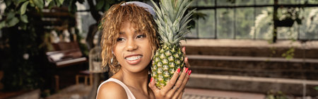 Smiling young african american woman with braces wearing headscarf and holding pineapple and...