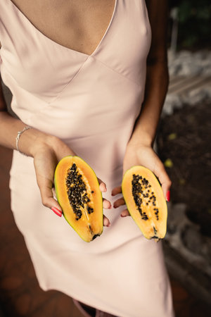 Cropped view of young african american woman in summer dress holding cut and fresh papaya and standing in blurred garden center, fashion-forward lady inspired by tropical plants, summer conceptの写真素材