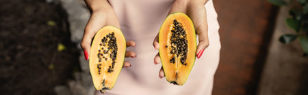 Cropped view of young african american woman in summer dress holding cut and fresh papaya while standing in blurred indoor garden, fashion-forward lady inspired by tropical plants, bannerの写真素材