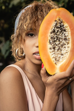 Portrait of young and fashionable african american woman in headscarf and summer dress holding cut and ripe papaya and covering face near plants, stylish lady blending fashion and natureの写真素材