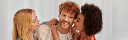 non traditional family, polygamy, three adults, happy interracial women hugging redhead man,...