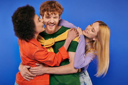 alternative relationships, polyamory lovers, cheerful multiethnic women kissing and hugging young redhead man on blue background, studio shot, vibrant colors, modern family, bonding and acceptanceの写真素材
