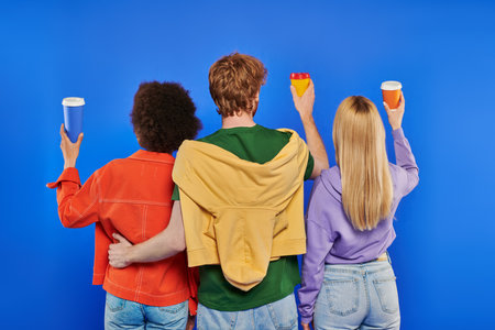love triangle, polyamory lovers, back view of young redhead man and multiracial women holding coffee to go on blue background, studio shot,  takeaway drink, vibrant colors, modern familyの写真素材