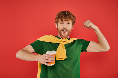 morning coffee, energy, showing muscle, redhead man with beard and curly hair holding paper cup on coral background, vibrant colors, male fashion, takeaway drink, male portrait, hot beverageの写真素材