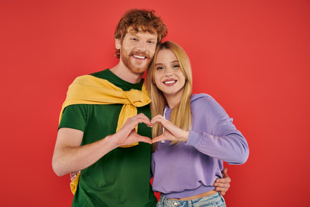 portrait of young couple looking at camera on coral background, showing heart sign with hands...
