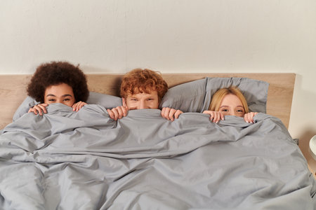 open relationship, polygamy, understanding, three adults having fun, hiding under blanket, redhead man and multicultural women, cultural diversity, acceptance, alternative familyの写真素材