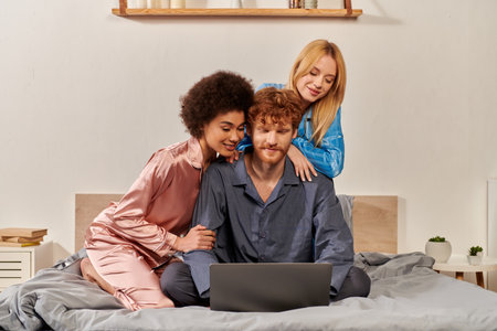 acceptance, open relationship, polygamy, understanding, three adults, redhead man and multicultural bisexual women in pajamas watching movie on laptop, bedroom, cultural diversityの写真素材