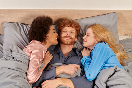 polyamory concept, three adults, happy man and interracial women in pajamas waking up together, morning, under blanket, bedroom, cultural diversity, bisexual, open relationship, polygamy, top viewの写真素材